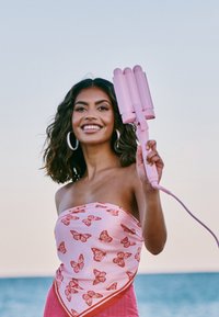 Różowa lokówka trójbarwna trzymana przez kobietę z falującymi włosami. Nosi różową bluzkę w motyle. Tło oceaniczne w delikatnych kolorach.