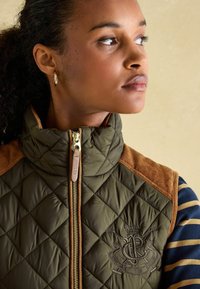 Prešívaná vestu olivovej farby s hnedými semišovými ramennými časťami a vyšitém logom, s vysokým golierom a zlatým zipsom.