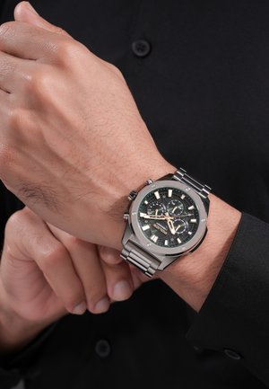 Muñeca izquierda con un reloj cronógrafo de metal plateado, esfera negra y manecillas doradas, sobre un fondo de camisa negra de manga larga.