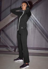 Schwarze Sportjacke mit hohem Kragen, reflektierendem Reißverschluss und enganliegenden Ärmelbündchen, kombiniert mit schwarzen Hosen und schwarzen Sportschuhen.