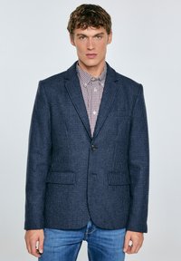 Mladý muž v námorníckej kárované blazer, pod ktorým má na sebe červenú a bielu károvanú košeľu a modré džínsy, stojí proti jednolitému pozadiu.