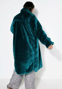Cappotto strutturato in peluche turchese con colletto a scialle, maniche lunghe e spacchi laterali; indossato su pantaloni argentati con paillettes, che mostrano un materiale morbido e testurizzato.