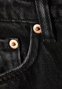 Primo piano di un denim nero con hardware in ottone e cuciture visibili. La texture appare liscia con una leggera lucentezza.