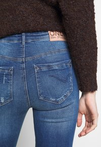 Pantalones vaqueros de denim azul con un ajuste de tiro medio, detalles desgastados y dos bolsillos traseros. La cinturilla presenta una etiqueta de cuero marrón claro.