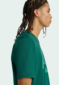 Zelené tričko s krátkym rukávom vyrobené z mäkkej látky, s okrúhlym výstrihom a grafickým potlačou na prednej strane. Povrch sa zdá byť hladký.