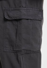 Donkergrijze cargobroek met een patchzak aan de zijkant, gestructureerde stof en zichtbare stikseldetails. Ontworpen voor functionaliteit en comfort.