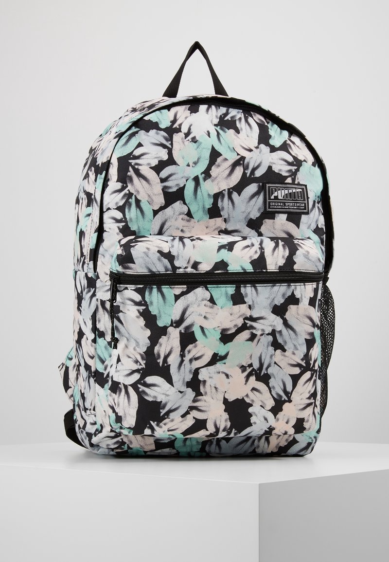 Sac à dos avec motif de feuilles noir, blanc, gris, beige et bleu sarcelle, poche avant zippée, poche latérale en mesh et boucle de transport supérieure sur surface blanche.
