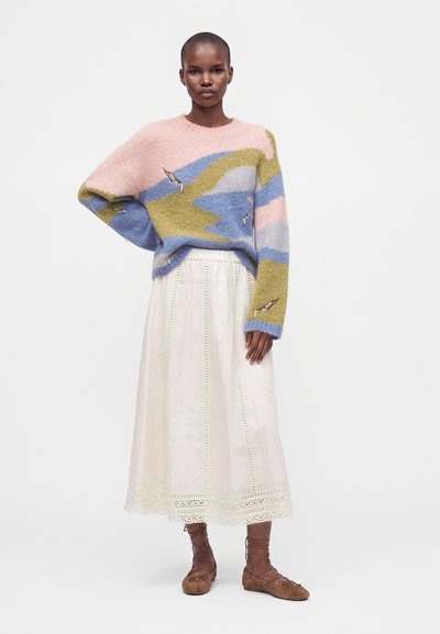 Femme portant un pull pastel à motif abstrait, une longue jupe blanche avec des détails œillets et des ballerines marron à lacets, debout sur un fond uni.
