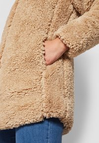 Hånd i lommen på en beige tekstureret teddyfrakke iført blå jeans, vist fra midt overkrop til midt lår mod en enkel baggrund.