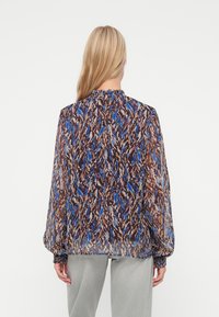 Mujer con cabello rubio largo que lleva una blusa de manga larga y cuello alto con un patrón abstracto en azul, marrón y beige, y pantalones gris claro, de espaldas.