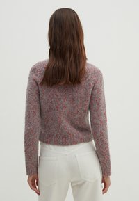Woman with long brown hair wearing a grey and red speckled fuzzy sweater and off-white pants, standing against a plain light background.