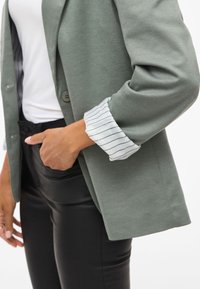 Lichtgroene blazer met twee knopen, voorzien van een wit gestreepte manchet. Gepaard met een getailleerde zwarte taille en elegante, glanzende zwarte broek.