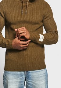 Man draagt een bruine geribbelde gebreide trui met een patch op de mouw en blauwe vervaagde jeans, handen ineengestrengeld bij de taille.