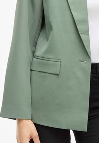 Blazer verde con un tessuto liscio e testurizzato. Presenta una tasca singola e un design a revers. Le maniche sono leggermente oversize.