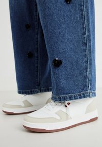 Sneakers bianchi e beige con dettagli perforati, abbinati a pantaloni in denim blu con decorazioni floreali nere.
