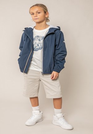 Jeune personne portant une veste à capuche bleu marine, un t-shirt blanc avec un motif graphique, un short beige, des chaussettes blanches et des baskets montantes blanches, debout les mains dans les poches.