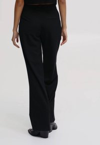 Pantalon noir à jambes larges en tissu léger, avec une texture lisse et un détail discret de poche arrière. Porté avec des chaussures noires.
