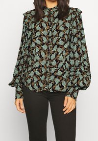 Blusa a maniche lunghe in tessuto scuro con motivo floreale verde, caratterizzata da spalle arricciate e chiusura con bottoni, abbinata a pantaloni neri.