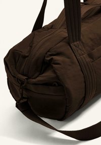 Hnědá taška přes rameno vyrobená z texturované tkaniny, zaobleného tvaru, s uzávěrem na zip, silnými popruhy a kovovým detailem na sponě.