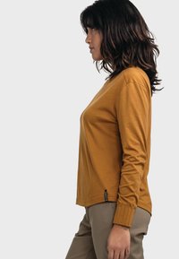 Langärmliges senfgelbes Oberteil aus weichem Stoff, mit einer lässigen Passform, gerippten Ärmelbündchen und einem seitlichen Logoetikett. Kombiniert mit hellen Hosen.