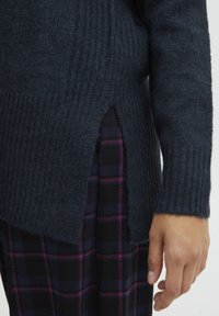 Ciemnoniebieski sweter dzianinowy z pionowym prążkowaniem, bocznymi rozcięciami i długimi rękawami. Połączony z czarną spódnicą w fioletową kratę.