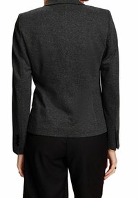 Kvinna iklädd en åtsittande mörkgrå texturerad kavaj och svarta byxor, sedd bakifrån, med händerna avslappnat vid sidorna.