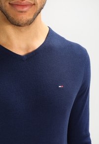 Man i en marinblå V-ringad tröja med en liten röd, vit och blå logotyp på vänster bröst mot en enkel ljus bakgrund.