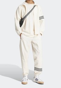 Light cream tracksuit with elastic cuffs and pocket. Features a zip-up jacket, a plain t-shirt, and matching pants. Black stripe accents.