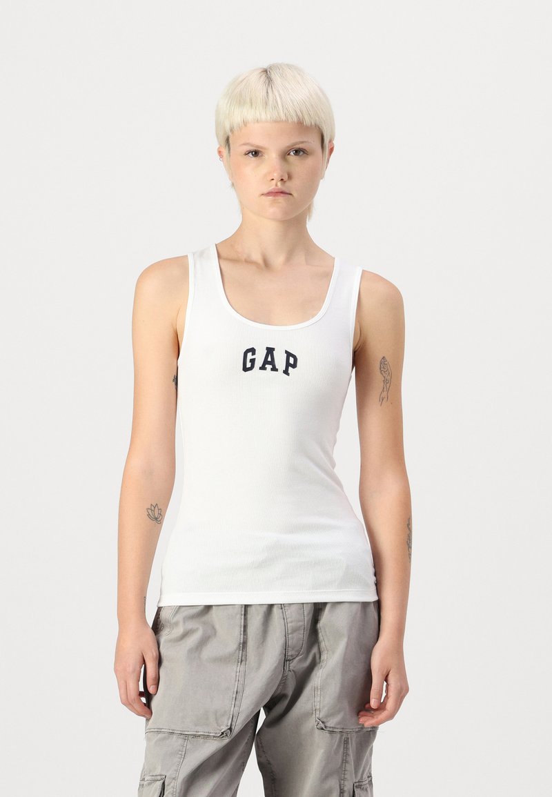 Camiseta sin mangas blanca con textura de canalé que presenta un logo en oscuro azul de "GAP" en el frontal. Diseñada con un escote redondeado y tirantes delgados.