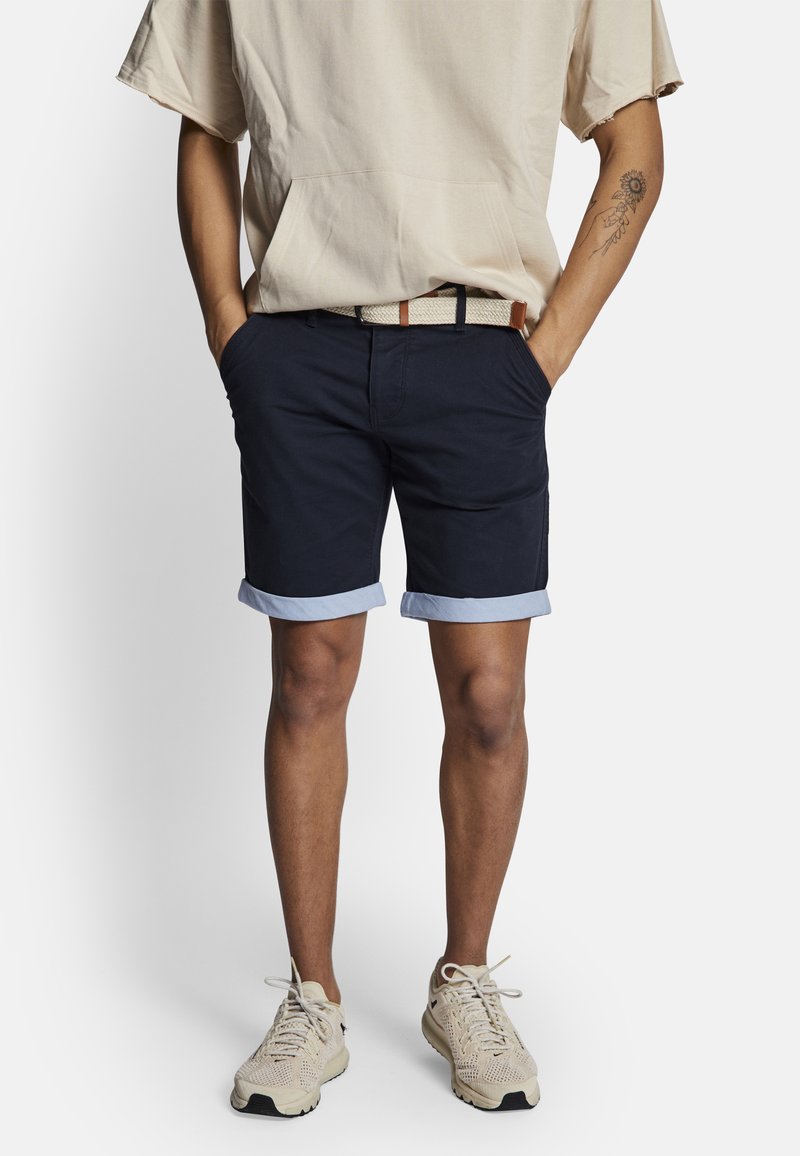 Shorts navy avec des revers bleu clair, en coton, dotés de poches latérales et d'une ceinture tissée avec un accent marron. Associés à des baskets beiges.