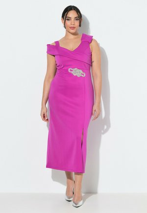 Kobieta w jasnoróżowej, bezrękawowej sukience z rozcięciem na udzie i ozdobną srebrną broszą, stojąca na jednolitym białym tle.