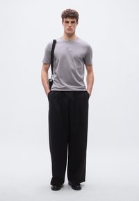 Γκρι t-shirt με στρογγυλή λαιμόκοψη και διακριτικό λογότυπο, συνδυασμένο με μαύρο παντελόνι φαρδιάς γραμμής και μαύρα παπούτσια, σε απλό φόντο.