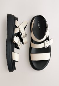 Stevige sandalen met een zwarte rubberen zool, dikke, crèmekleurige neplederen bandjes en twee verstelbare zilveren gespen.