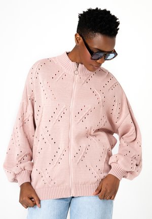 Osoba nosząca przesadnie duży jasnoróżowy sweter zapinany na zamek z wzorem dziur, jasnoniebieskie dżinsy i ciemne okulary przeciwsłoneczne, patrząca w dół.