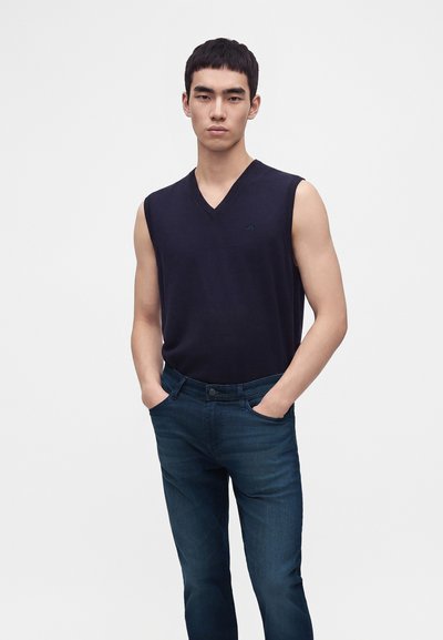 Jeune homme portant un gilet en tricot bleu marine sans manches et un jean bleu foncé, les mains dans les poches, sur un fond clair uni.