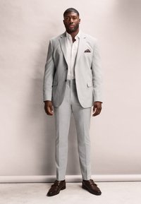 Completo grigio chiaro con design a uno petto, abbinato a una camicia bianca e a un fazzoletto da taschino a fantasia. Scarpe in pelle marrone completano il look.