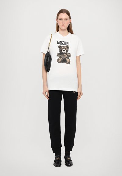 Hvit bomull t-skjorte med svart bjørneillustrasjon og "MOSCHINO" tekst, sammen med svarte joggebukser og svarte slip-on sko. Langt rett hår.