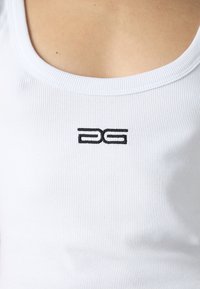 Närbild av en vit rippad linne med en liten svart broderad logotyp centrerad under halsringningen.