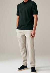 Ciemnozielona koszulka polo z małym złotym logo, zestawiona z jasnobeżowymi spodniami i czarnymi trampkami. Materiał wydaje się gładki, o klasycznym kroju.