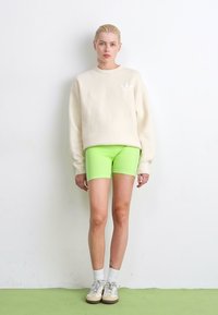 Lichtbeige sweatshirt met een klein wit logo, gecombineerd met felgroene shorts, witte sokken en beige sneakers. Eenvoudig en modern ontwerp.