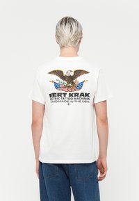 White cotton t-shirt featuring a colorful eagle graphic on the back, text reading "KRAK Electric Tattoo Machines," and short sleeves.