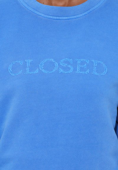 Μπλε φούτερ με στρογγυλή λαιμόκοψη και μακρύ μανίκια. Φέρει την επιγραφή "CLOSED" κεντημένη με ανοιχτό μπλε νήμα διαγωνίως στο στήθος.