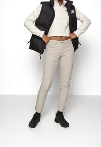 Chaleco puffy negro sobre una parte superior de forro polar blanca con cremallera de un cuarto, combinado con pantalones beige claros de corte slim y zapatillas deportivas negras. Diseño simple, sin patrones.