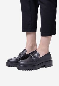 Scarpe slip-on in pelle nera con suola spessa, punta arrotondata, dettagli decorativi e una superficie liscia. Indossate con pantaloni neri a 3/4.