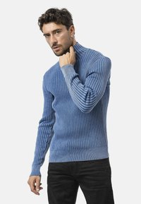 Man in een blauwe geribbelde coltrui die met zijn rechterhand de kraag aanpast, staande tegen een effen lichte achtergrond.