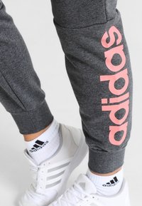 Grijze joggers met een roze "adidas" logo langs het linkerpijp, gecombineerd met witte sneakers en gestreepte sokken. Het materiaal lijkt zacht en textuur te hebben.