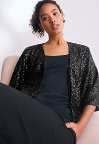 Veste noire à sequins avec une coupe ample et manches courtes, portée sur un haut sombre, sur un fond clair. Texture lisse.