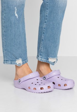 Fødder iført lyse lilla slip-on træsko med ventilationshuller, parret med blå revnede denimjeans mod en ensfarvet grå baggrund.