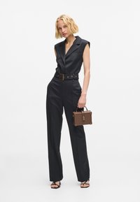 Mustaga kohandatud üldkukkuja, millel on istuv talje ja laiad püksid. Omab kraed, metallist kinnitusega vööd ja pruuni struktureeritud käekotti.