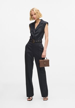 Čierny prispôsobený overal s priliehavým pásom a širokými nohavicami. Obsahuje golier, opasok s kovovou sponou a hnedú štruktúrovanú kabelku.